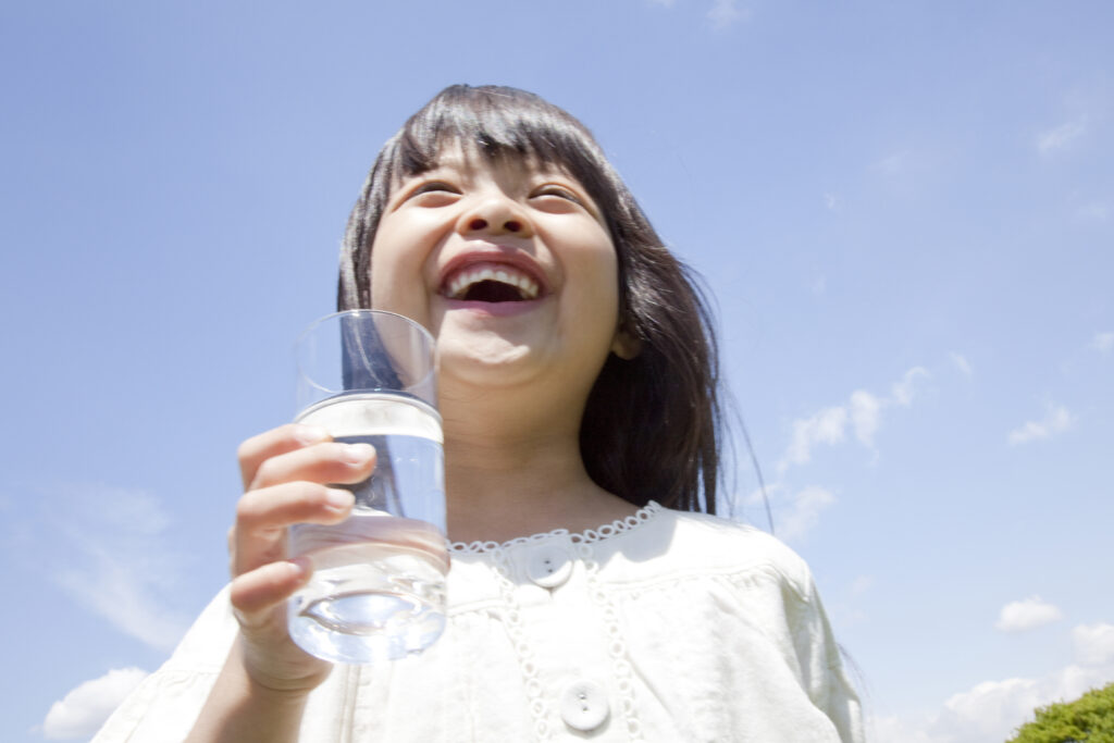 白い服を着た女の子が水の入ったガラスのコップを片手に、青空の下で笑っている様子