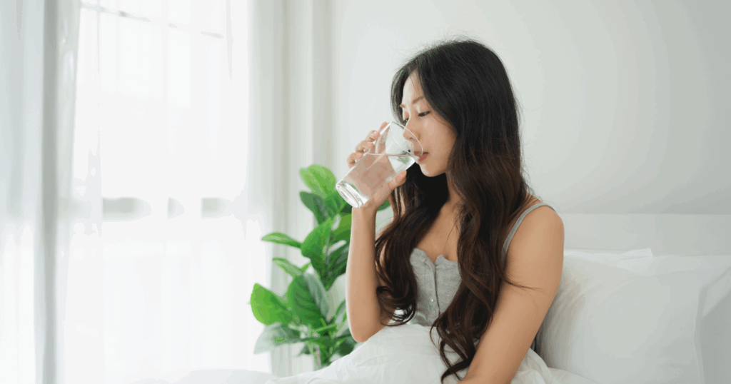朝ベッドに入りながら、ガラスのコップに入れた水を飲む女性の写真