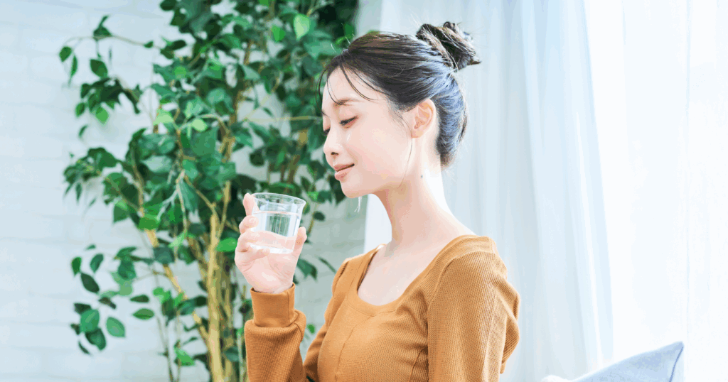 部屋の中で水の入ったガラスのコップを片手に持ち微笑む女性の写真