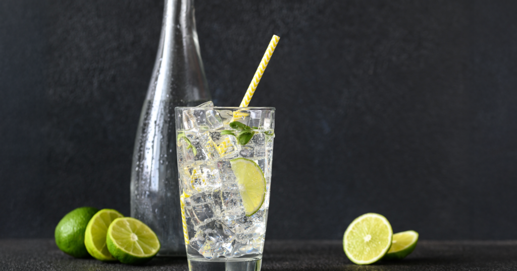 ブラックの背景の前に置かれた、ガラスのコップに入ったライム入り炭酸水。ガラスのボトルもコップの後ろにある。