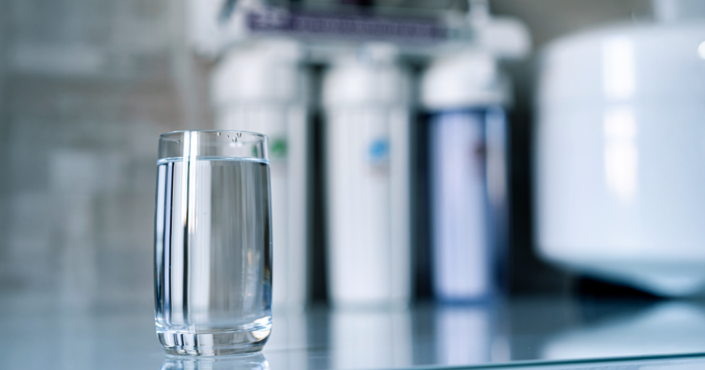 背景に逆浸透膜浄水器のフィルターがあり、手前に水の入ったグラスが置いてある画像