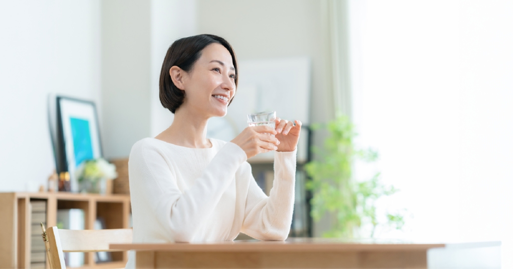 女性が家のテーブルに肘をつき水の入ったグラスを持っている様子。