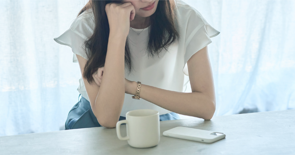 マグカップの置かれたテーブルに肘をついて座る女性。生理痛でしんどそうな雰囲気。