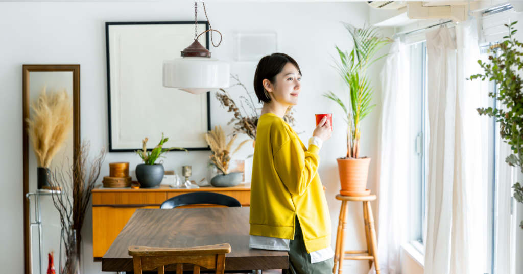 部屋で女性がマグカップを片手にダイニングテーブルへもたれかかっている様子