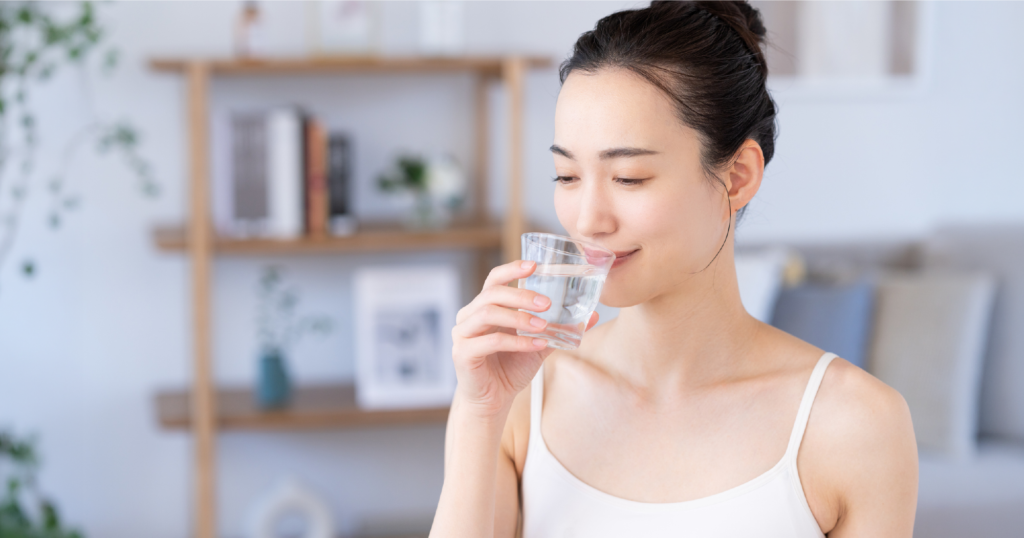 キャミソール姿の女性がガラスのコップに入った水を飲もうとしているシーン。