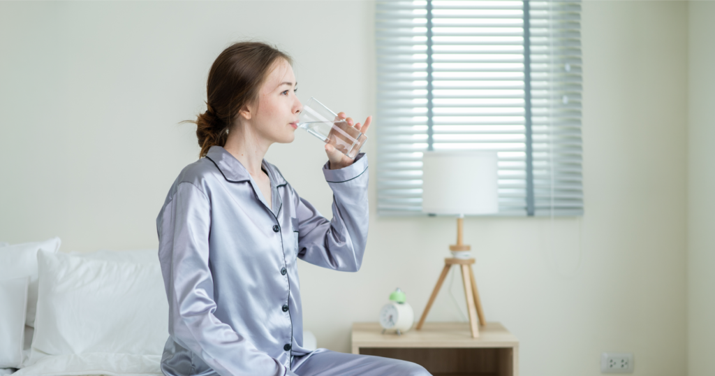 ベッドに座りガラスのコップから水分補給をしている女性。