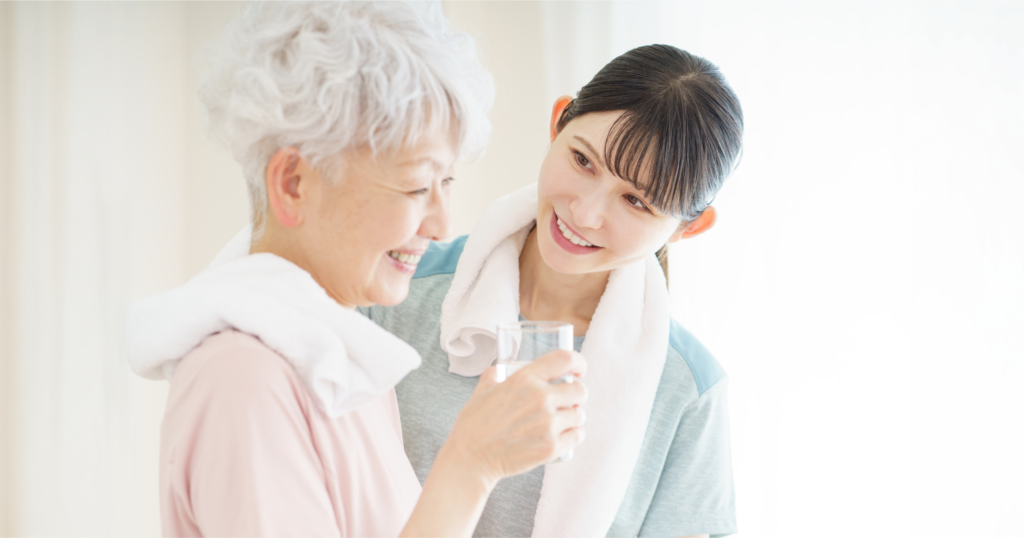 ガラスのコップに入った水をもつ高齢女性と、高齢女性をサポートする若い女性。