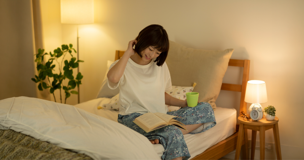 ベッドに座りマグカップで水分補給をしながら本を読んでいる女性。