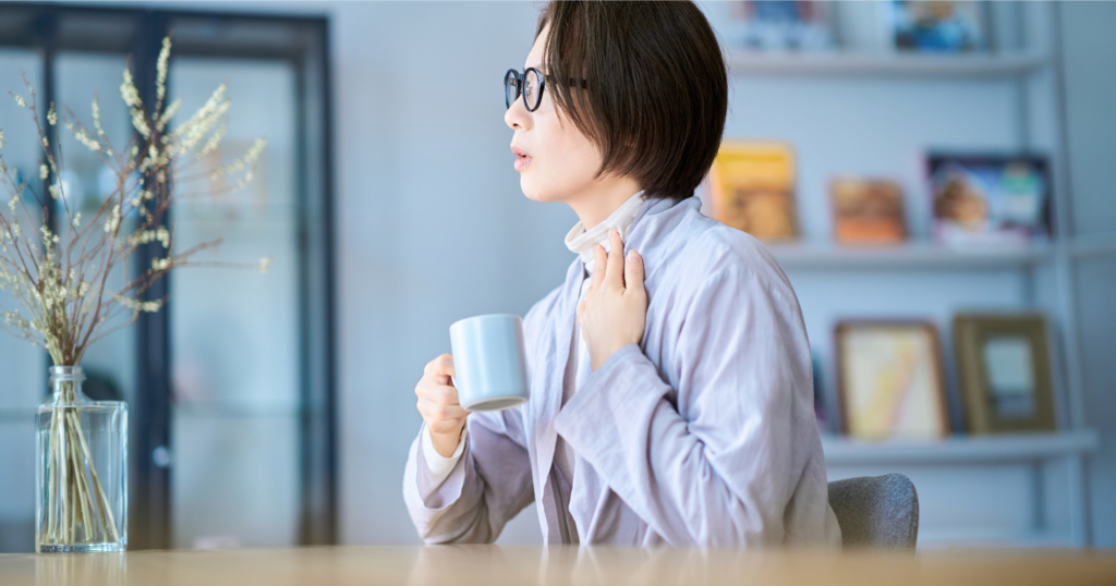 女性が喉に手を当てながら、もう片方の手でマグカップを持っている。リビングの机に座っている。